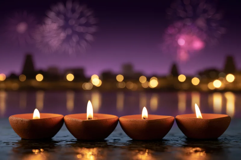 Four clay oil lamps with flickering flames floating on water at night, city skyline and fireworks in background