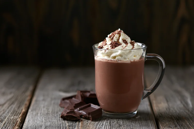 Glass mug of hot chocolate topped with whipped cream and chocolate pieces on a wooden table