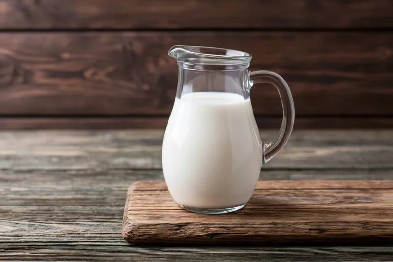 Glass pitcher filled with white milk on a wooden cutting board against rustic wood background