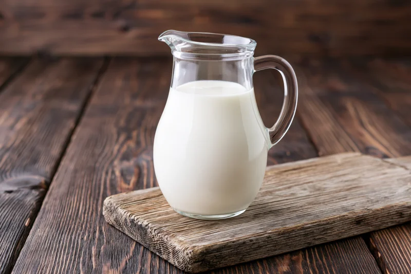 Glass pitcher filled with white milk on a wooden cutting board on a rustic table