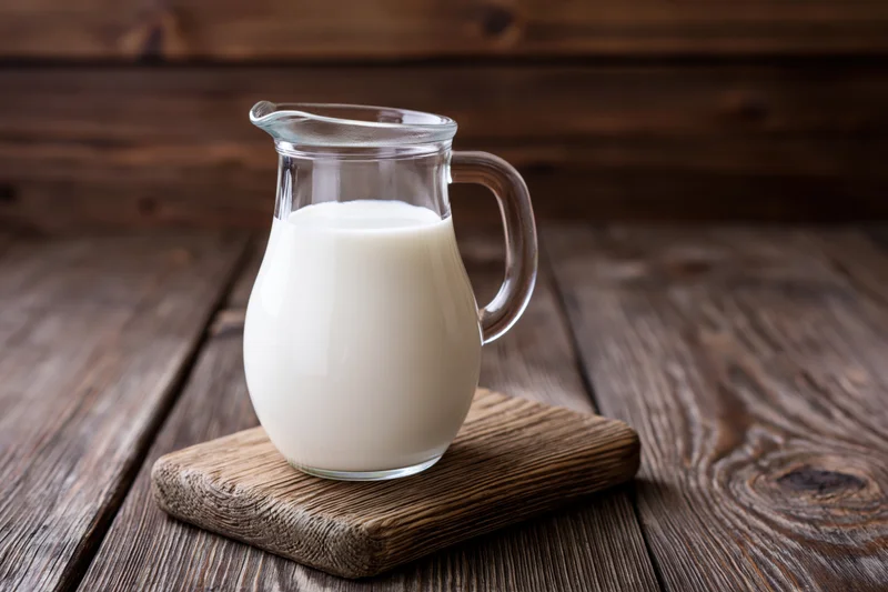 Glass pitcher filled with fresh white milk on a wooden cutting board atop a rustic wooden table
