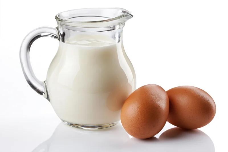 Glass pitcher filled with milk beside two brown eggs on white surface