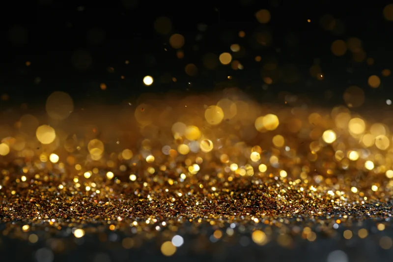 Gold glitter particles scattered across a dark surface with bokeh lights in the background