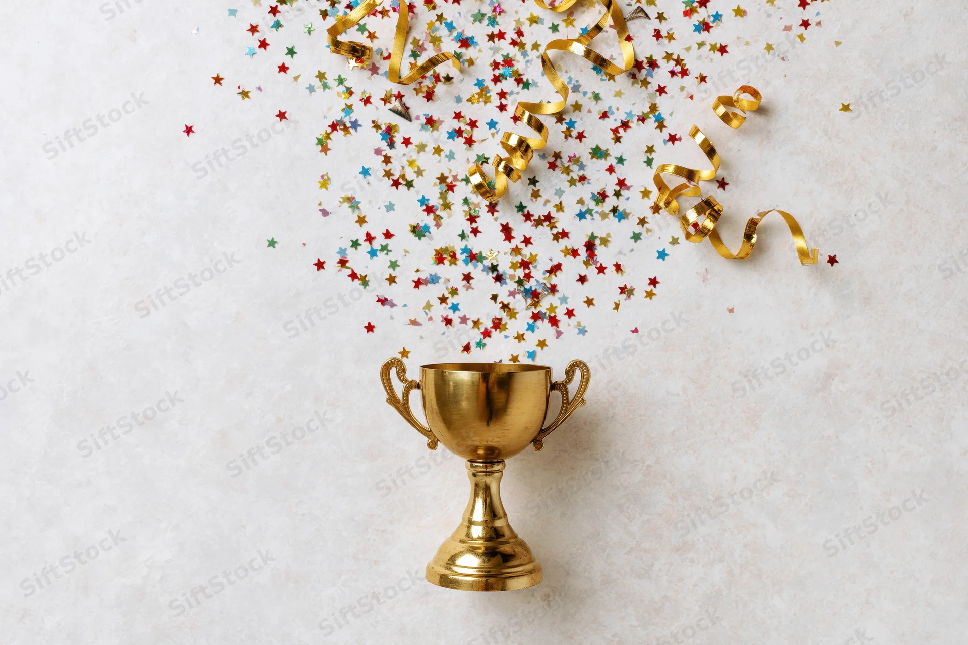 Gold trophy cup with colorful star confetti and golden ribbons falling above it on light surface