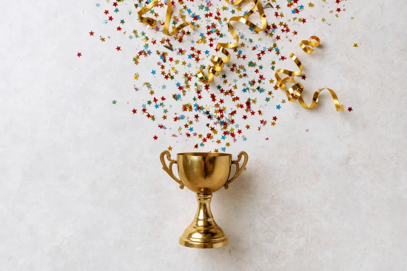 Gold trophy cup with colorful star confetti and golden ribbons falling above it on light surface