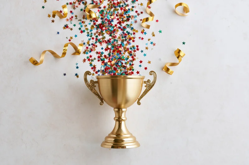 Gold trophy cup overflowing with colorful star confetti and golden ribbons on white surface
