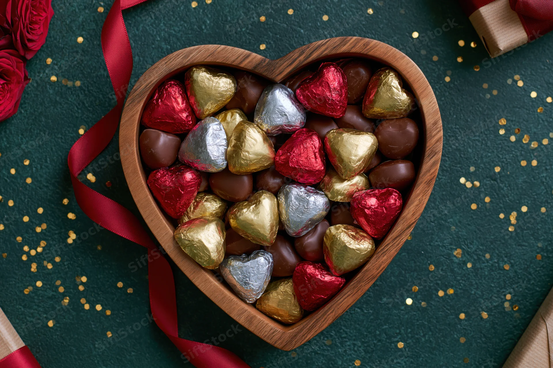 Heart-shaped wooden box filled with foil-wrapped chocolates on dark green surface with roses and ribbon