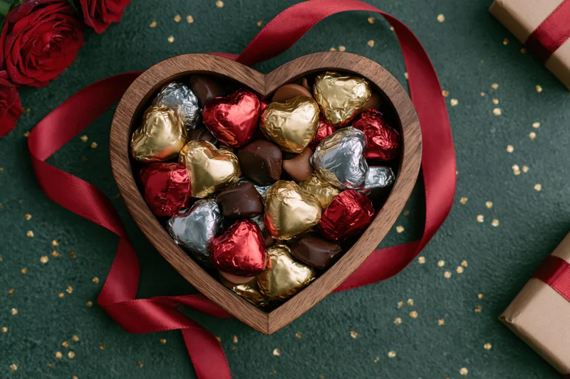 Heart-shaped wooden box filled with foil-wrapped chocolates on dark green surface with red ribbon and roses