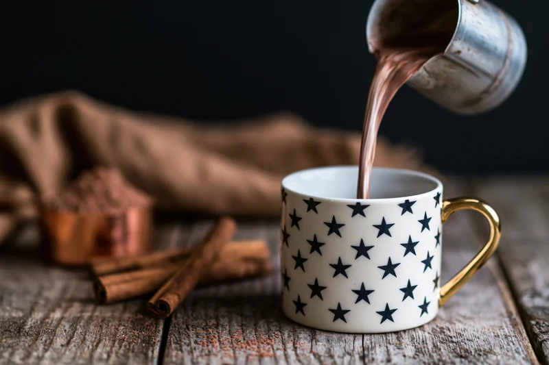 Hot cocoa being poured from a metal pitcher into a star-patterned mug on a wooden table