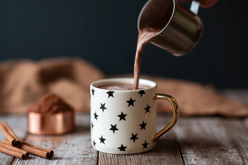 Hot cocoa being poured from a metal pitcher into a white star-patterned mug on a wooden table