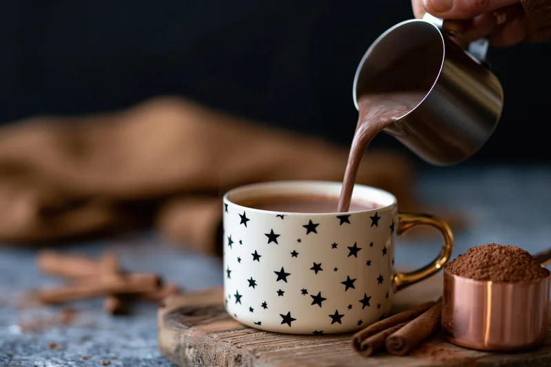 Hot cocoa being poured from a metal pitcher into a star-patterned mug on a wooden board with cinnamon sticks and a copper container nearby