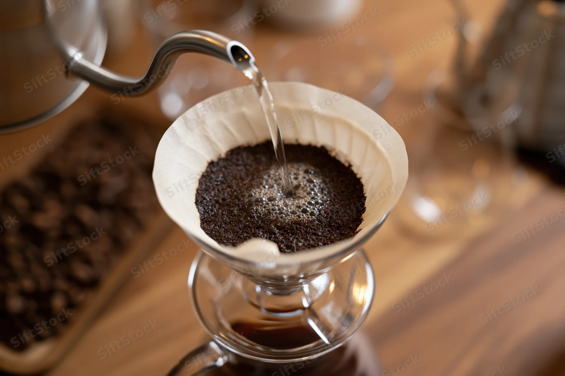 Hot water pouring from a gooseneck kettle into a paper filter filled with coffee grounds over a glass carafe on a wooden table