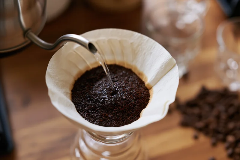 Hot water pouring from a gooseneck kettle into a paper filter filled with coffee grounds on a wooden table