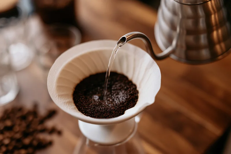 Hot water poured from a gooseneck kettle into a paper filter over coffee grounds on a wooden table