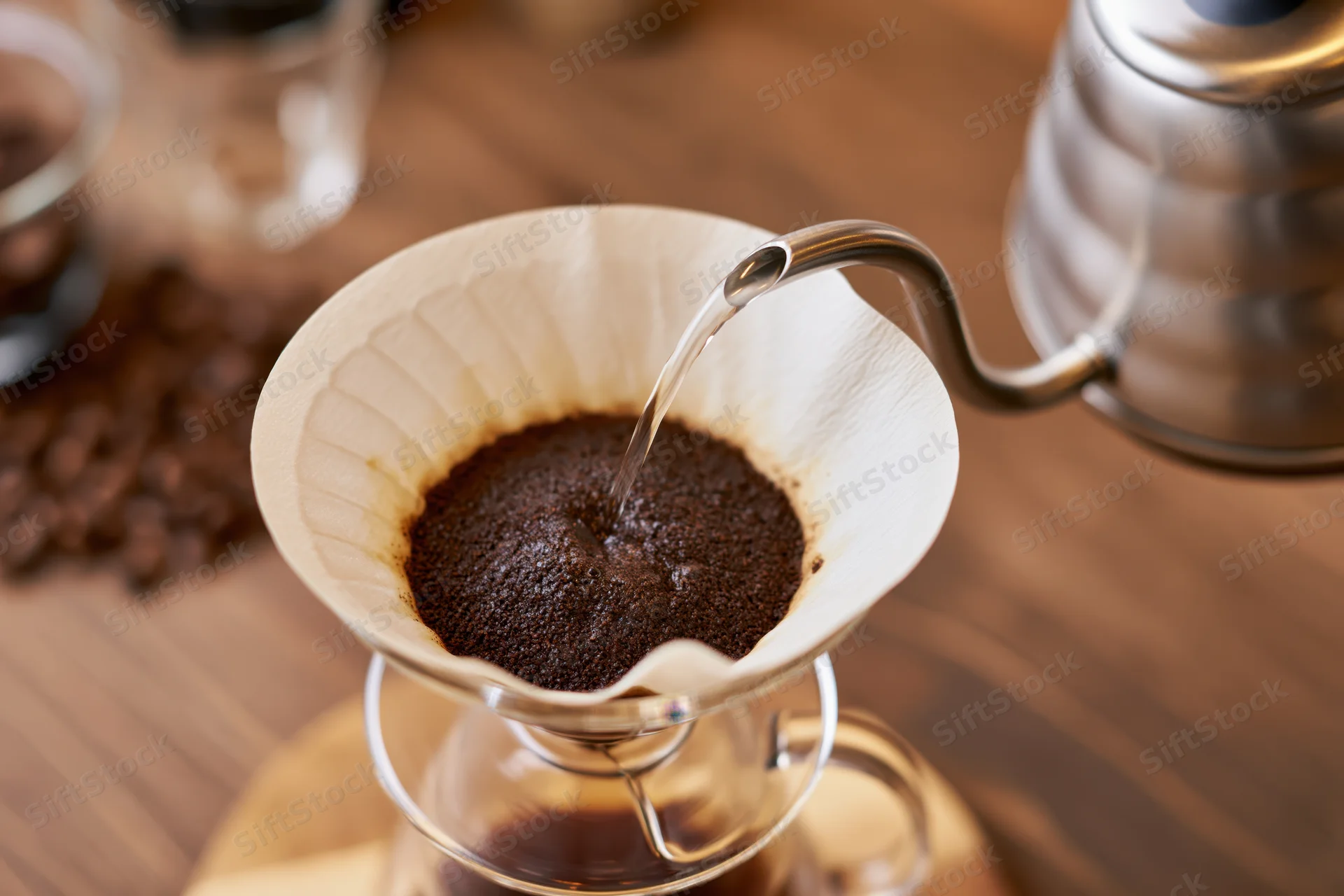 Hot water pouring from a gooseneck kettle into a paper filter filled with coffee grounds over a glass carafe on a wooden table