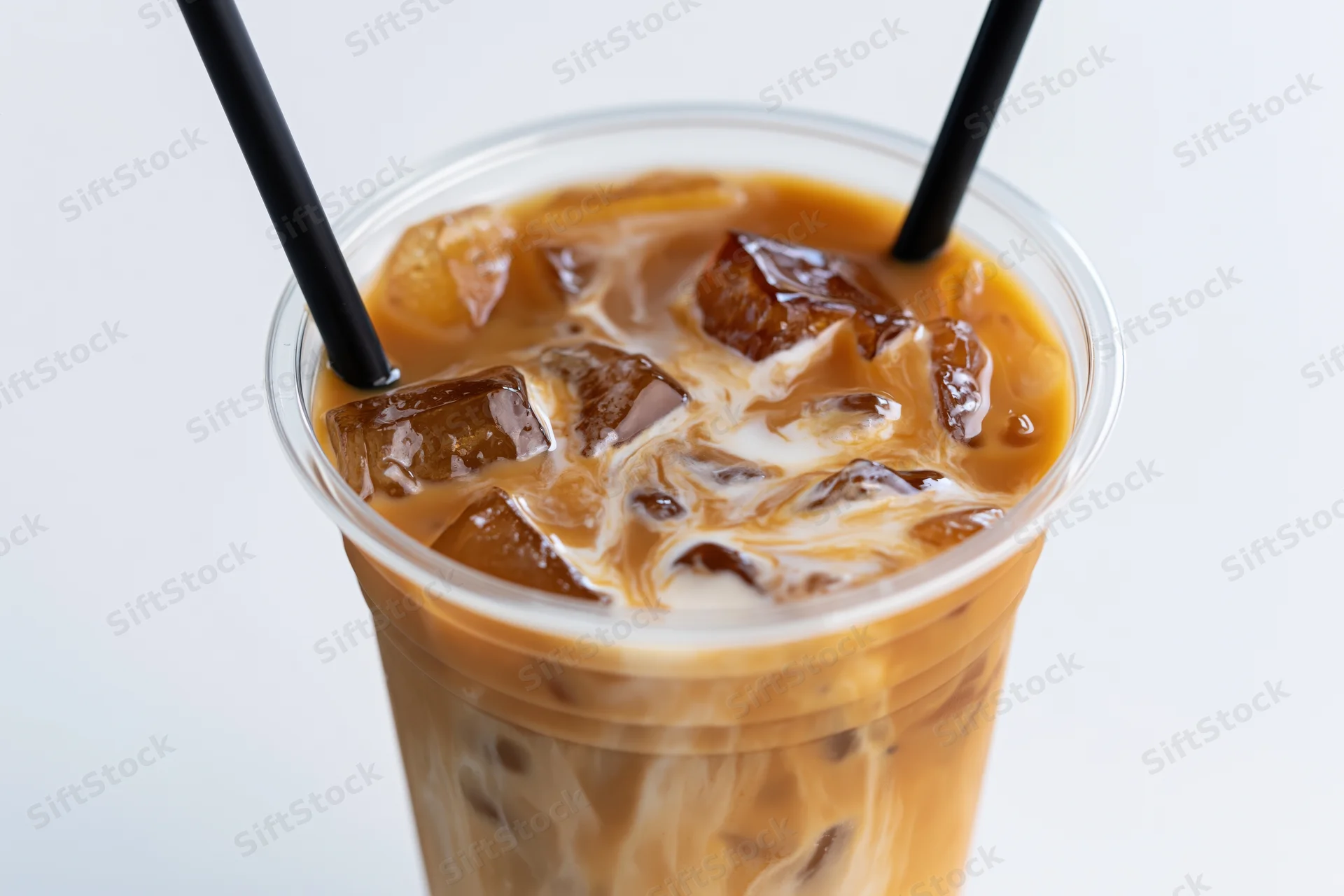 Iced coffee with cream swirling through ice cubes in a clear plastic cup with two black straws against white background