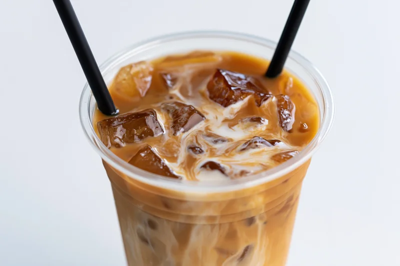 Iced coffee with cream swirling through ice cubes in a clear plastic cup with two black straws against white background
