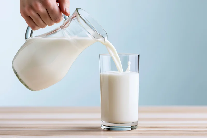 Hand pouring milk from a glass pitcher into a tall glass on a wooden table