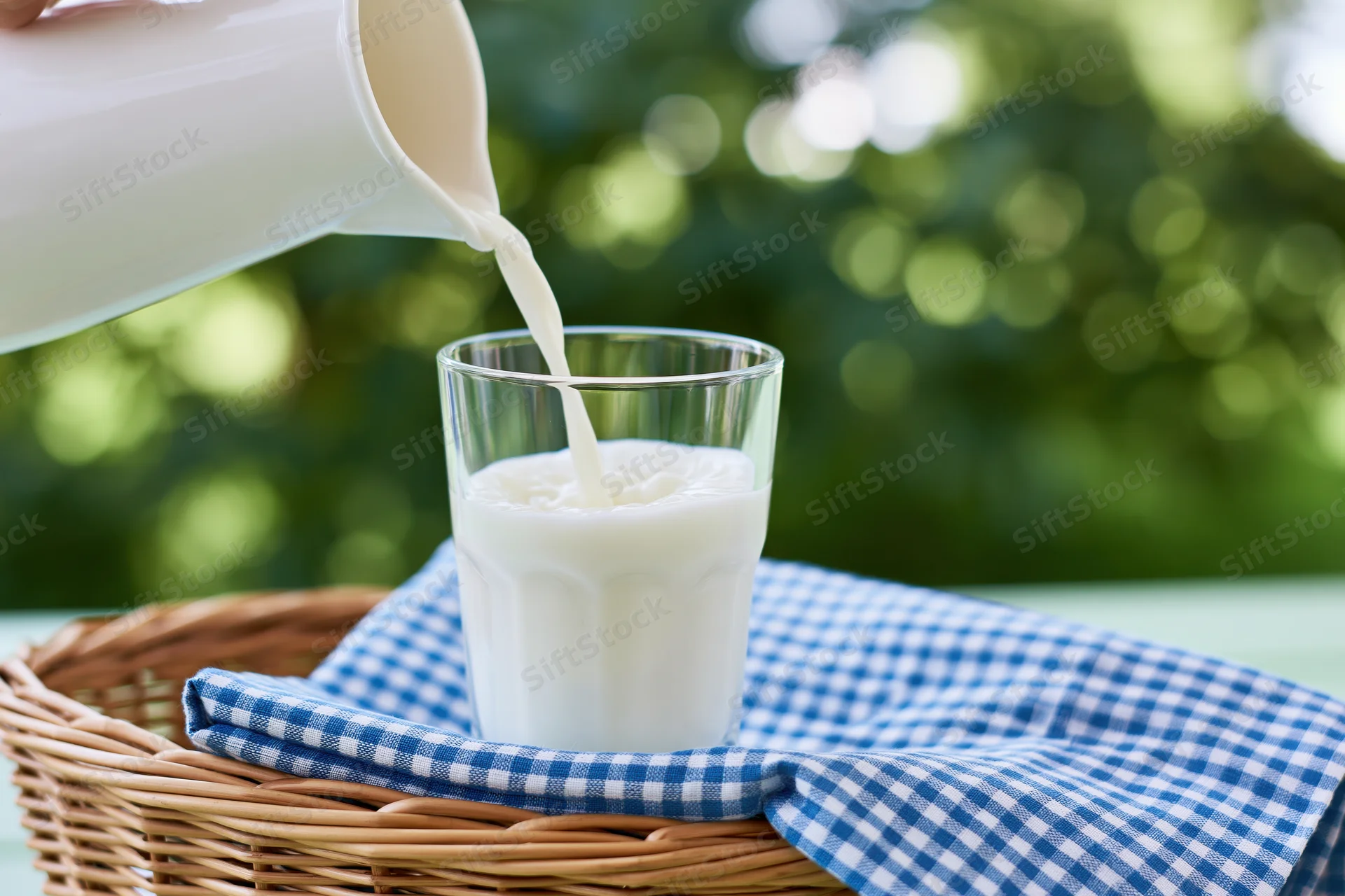 Milk being poured from a white jug into a clear glass on a checkered cloth in a wicker basket outdoors