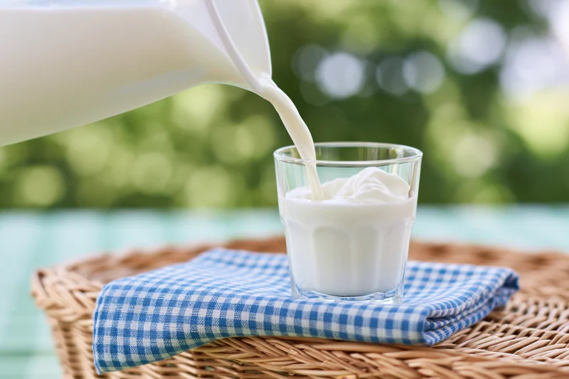 Milk being poured from a white jug into a clear glass on a checkered napkin in a wicker basket outdoors