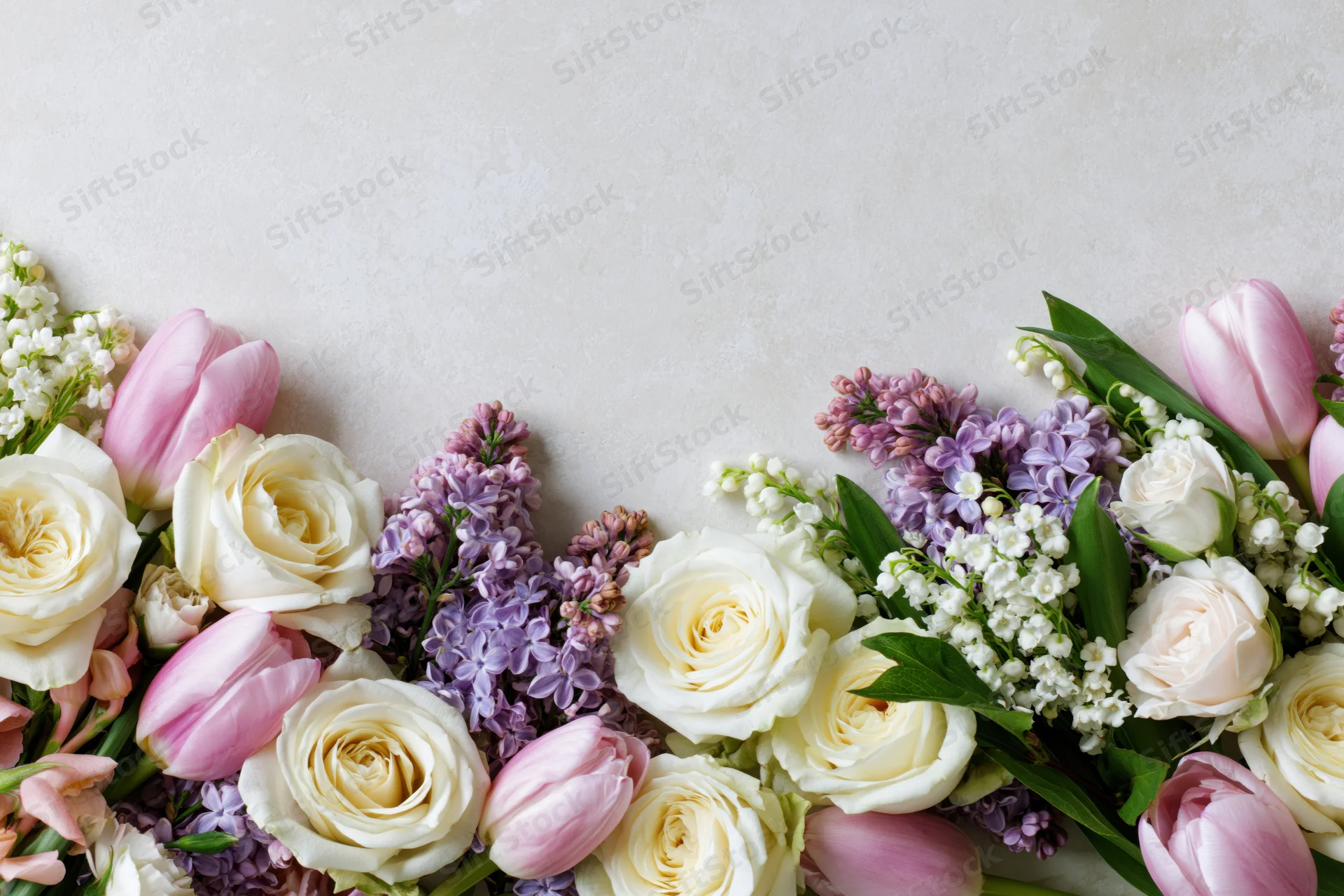 Spring bouquet of roses, tulips, lilacs and lily of the valley arranged along bottom edge on light surface