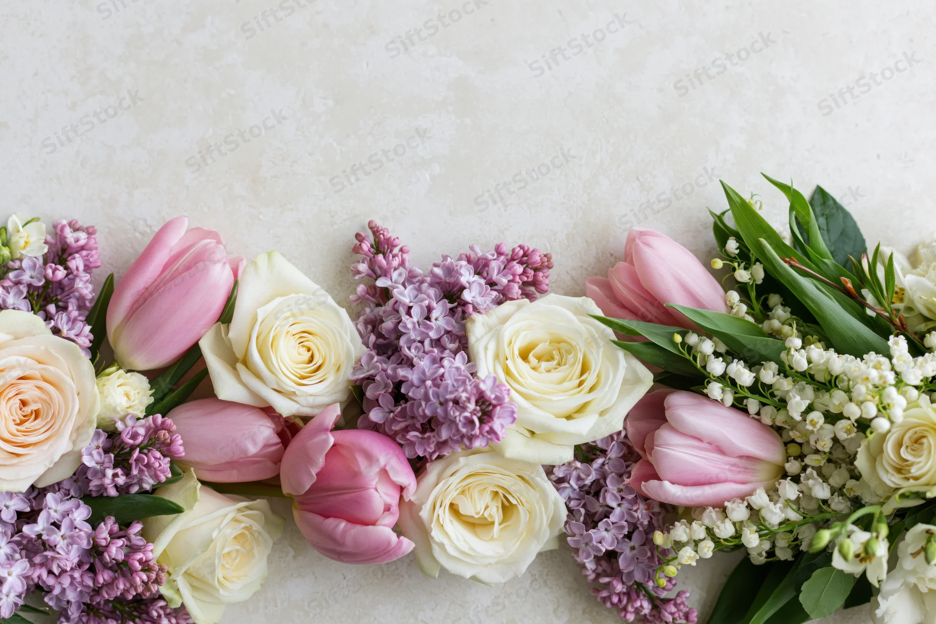 Spring bouquet of roses, tulips, lilacs and lily of the valley arranged along bottom edge on light textured surface