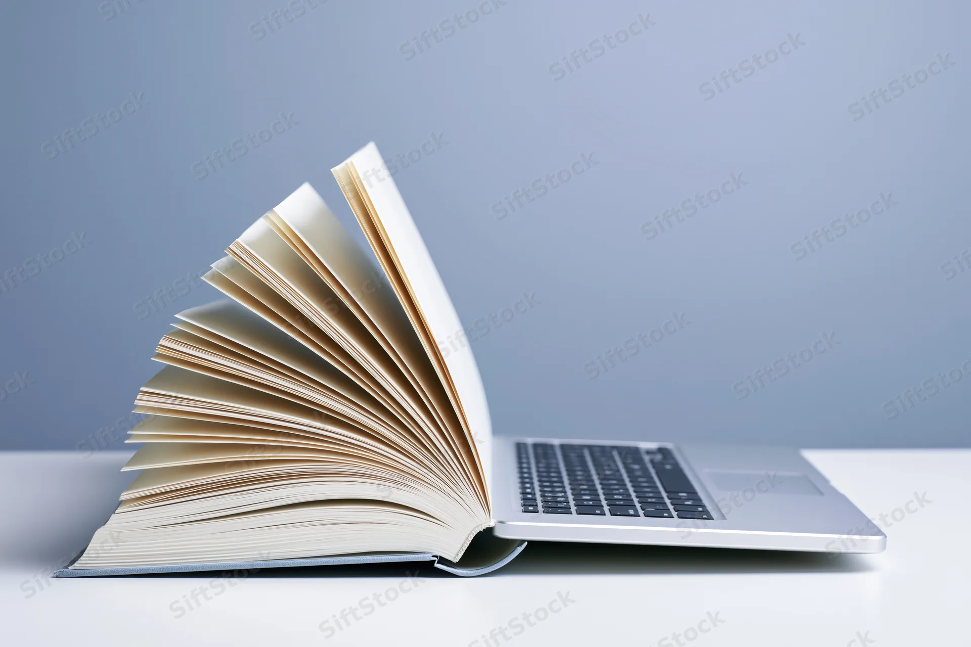 Open hardcover book with fanned pages merging into laptop on white desk