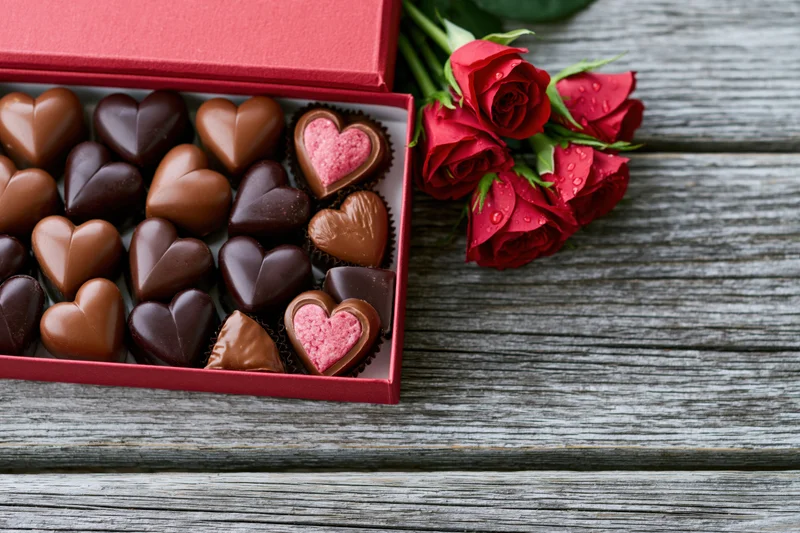 Open red gift box with heart-shaped chocolates and red roses on wooden surface