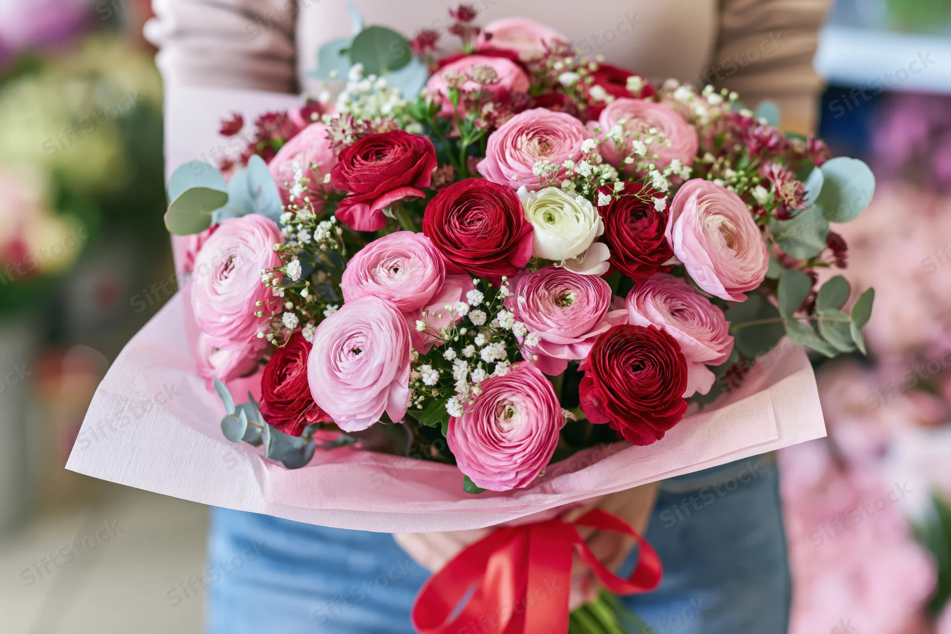Woman holding a lush bouquet of pink and red ranunculus flowers wrapped in pink paper with a red ribbon