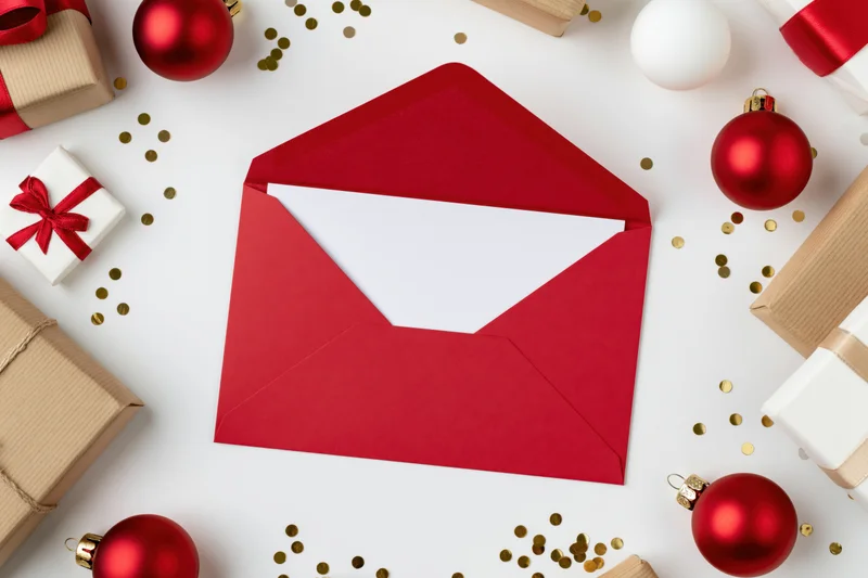 Open red envelope with blank white card inside surrounded by christmas ornaments and gifts on white surface