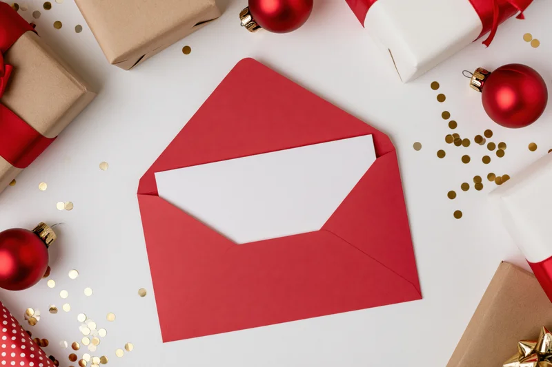 Open red envelope with blank white card inside surrounded by christmas gifts and ornaments on white surface