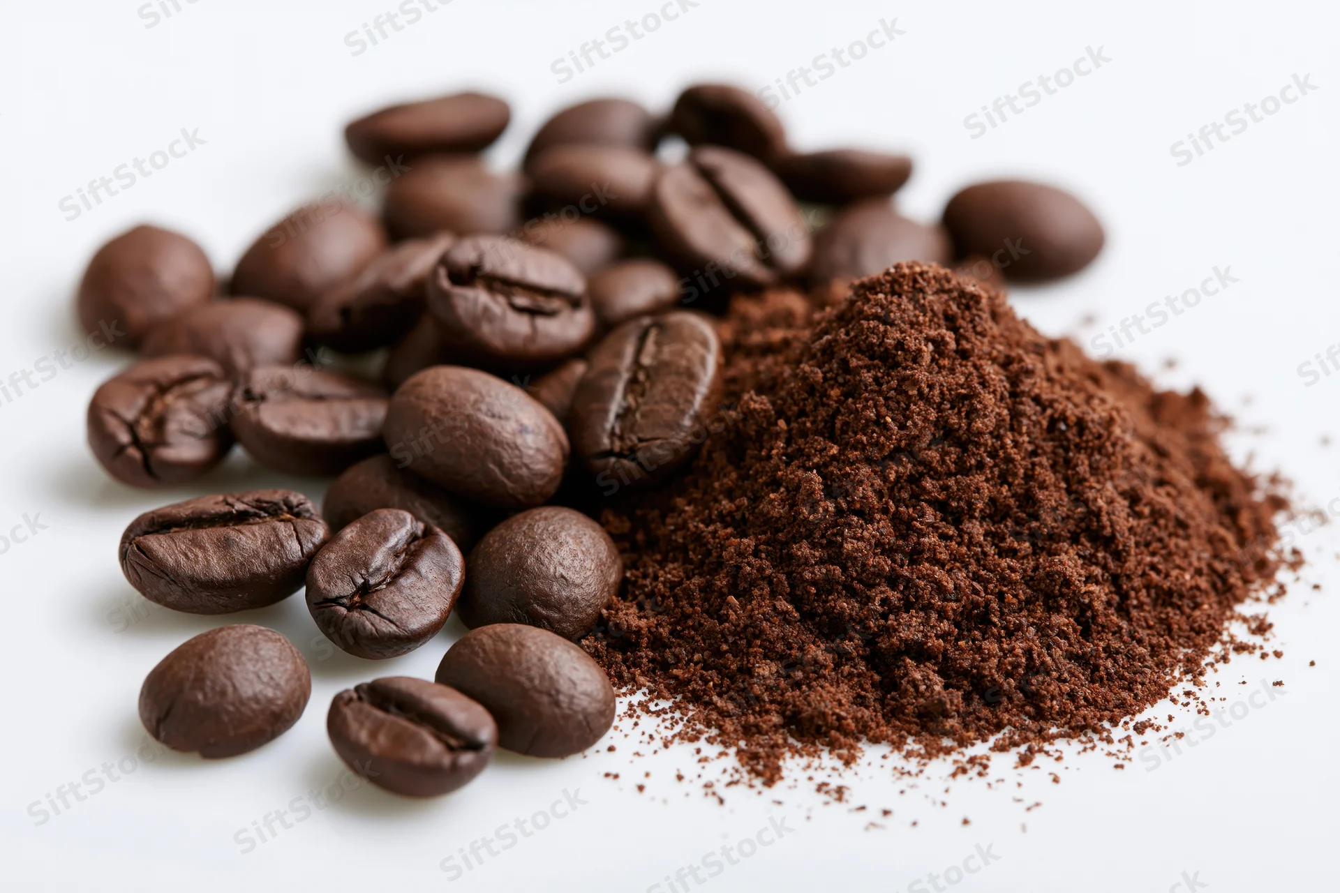 Scattered whole roasted coffee beans beside a small mound of ground coffee on white surface