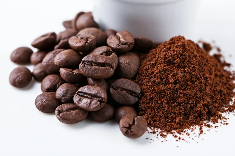 Whole roasted coffee beans and ground coffee piled beside a white cup on a white surface
