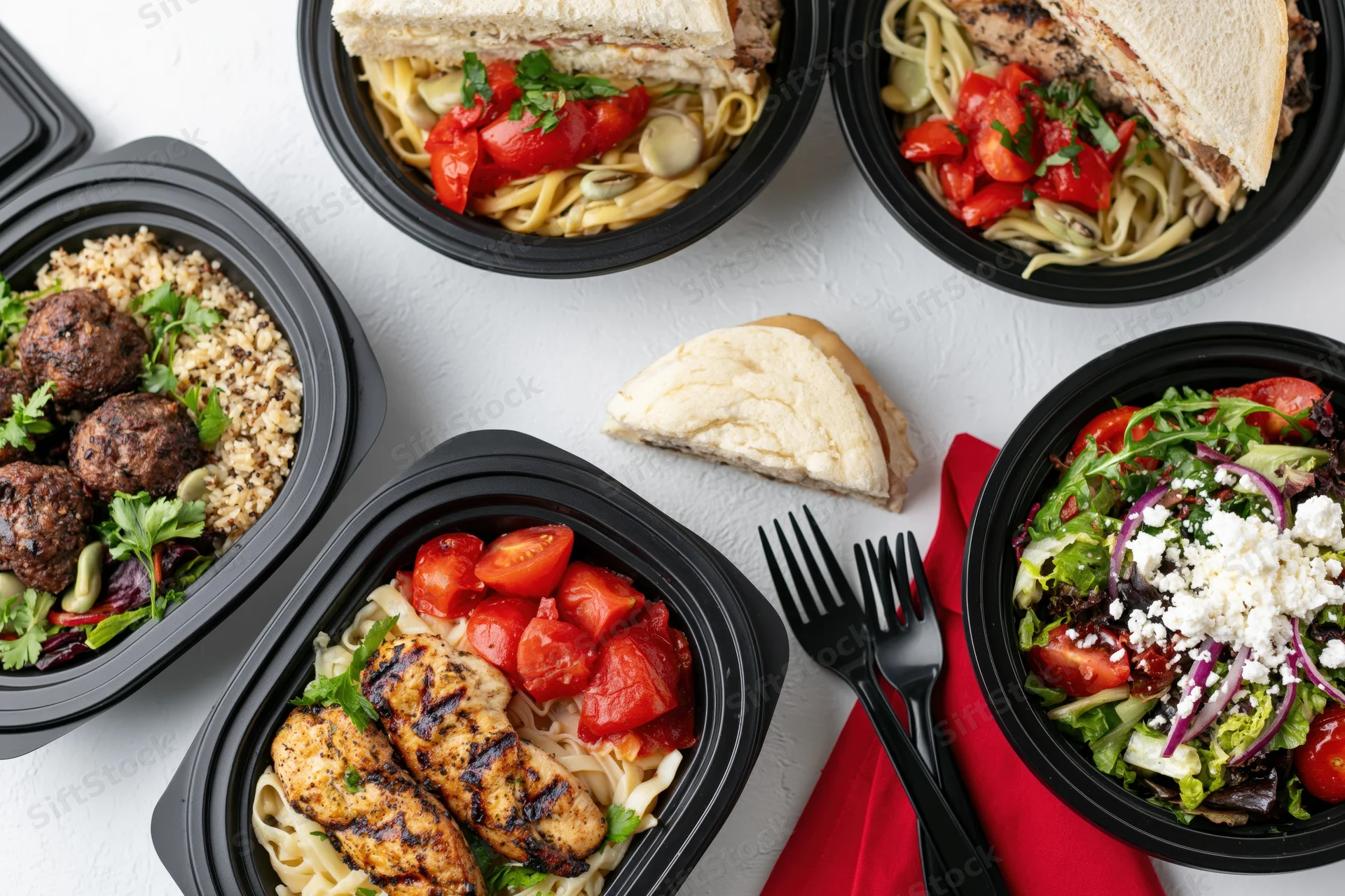 Several black plastic takeout containers filled with prepared meals arranged on a white surface with forks and a red napkin