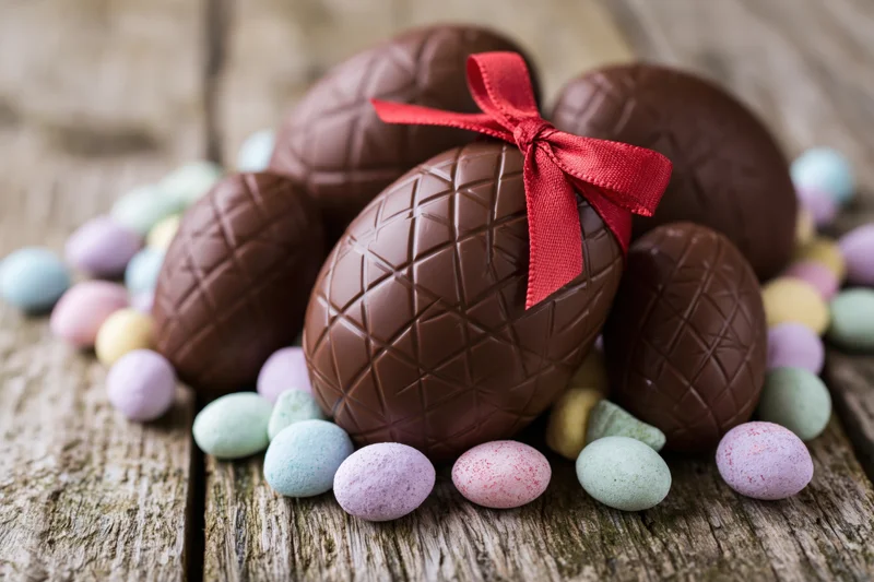 Chocolate easter eggs with red ribbon arranged on rustic wooden surface surrounded by pastel candy-coated eggs