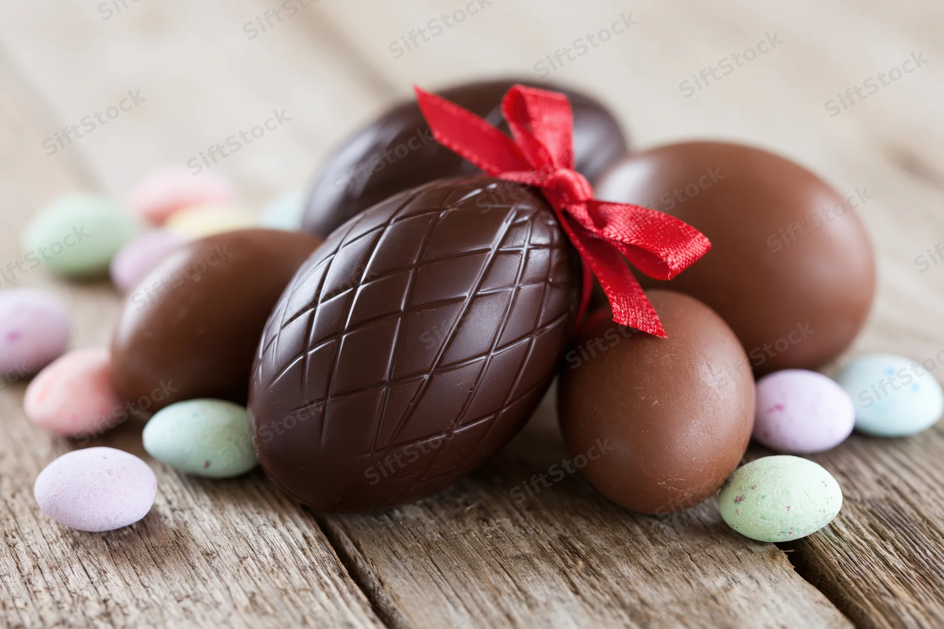 Chocolate easter eggs of various sizes arranged on a wooden table with a red ribbon and pastel candies around