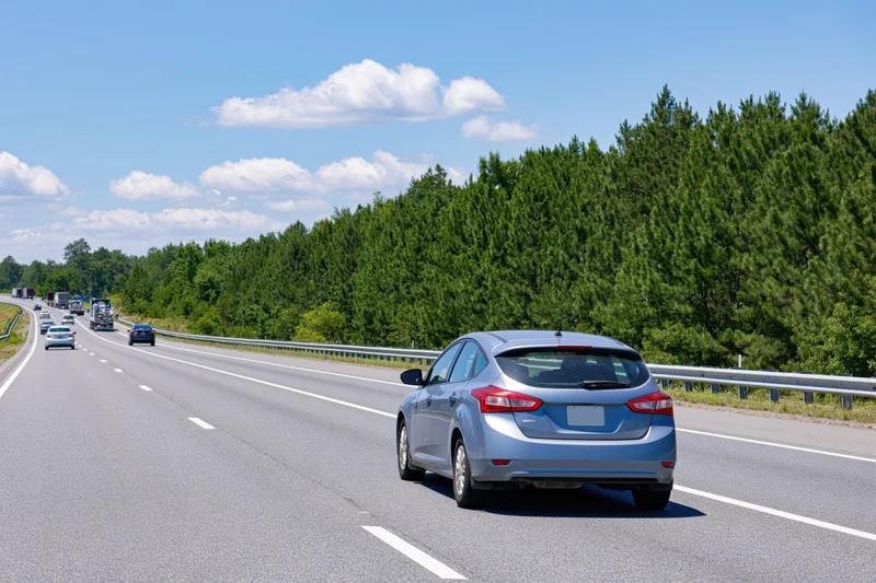 Silver hatchback driving away on a multi-lane highway bordered by trees under a blue sky