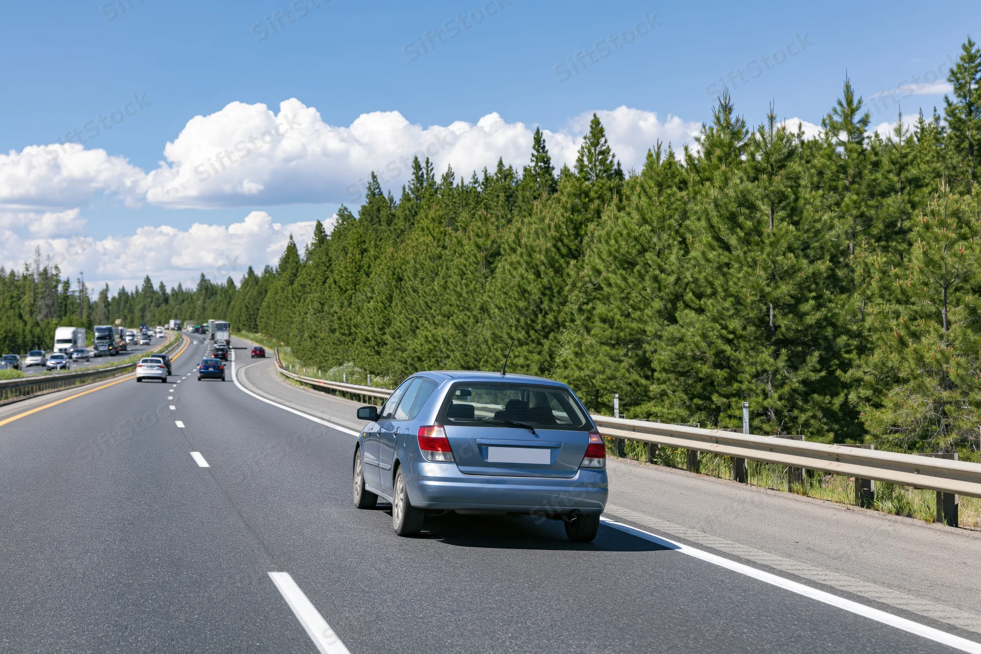 Silver hatchback driving away on a two-lane highway lined with pine trees under a blue sky with clouds
