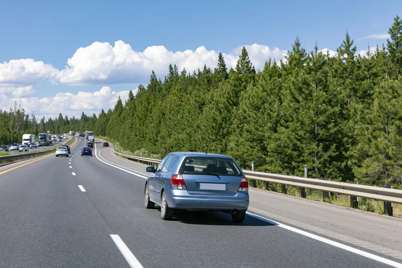 Silver hatchback driving away on a two-lane highway lined with pine trees under a blue sky with clouds