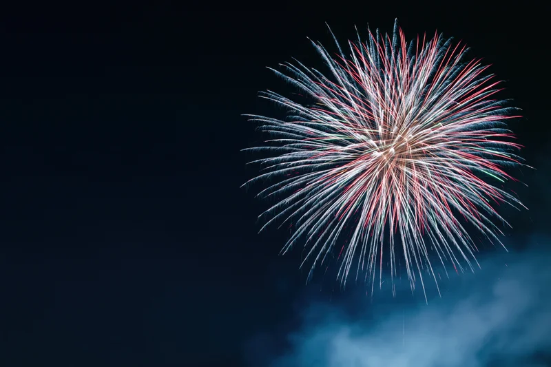 Large firework burst exploding in the night sky with red white and blue sparks and smoke below