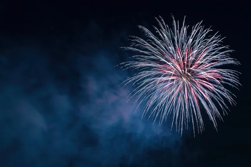 Large firework burst exploding in the upper-right of the dark night sky with smoke and colorful trails