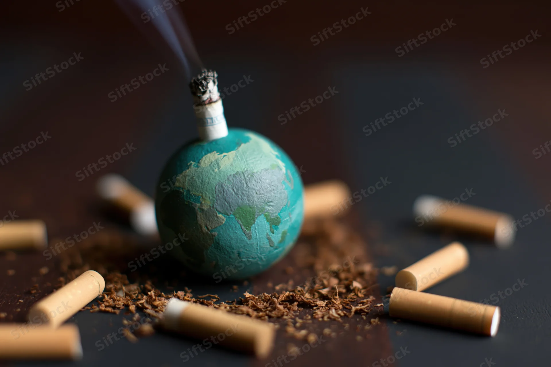 Small painted globe with lit cigarette inserted into top, surrounded by scattered cigarette butts and ash on dark surface