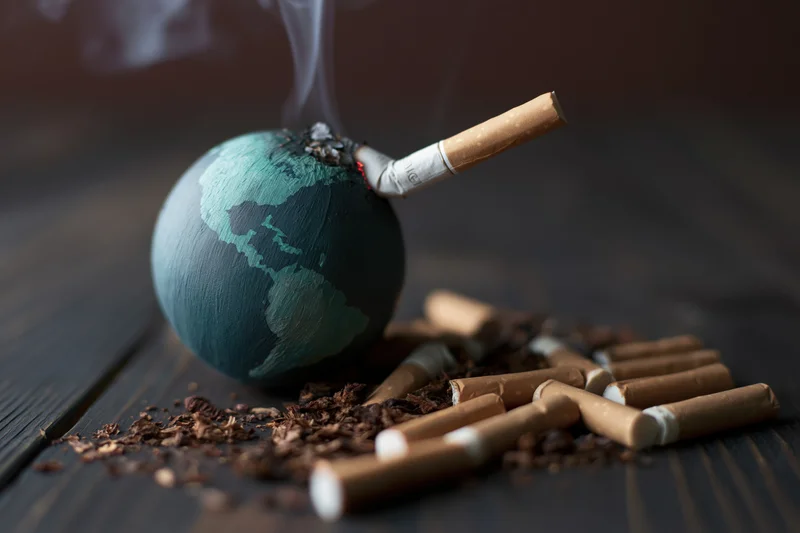 Painted globe with lit cigarette inserted into it, surrounded by cigarette butts and ash on dark wooden surface