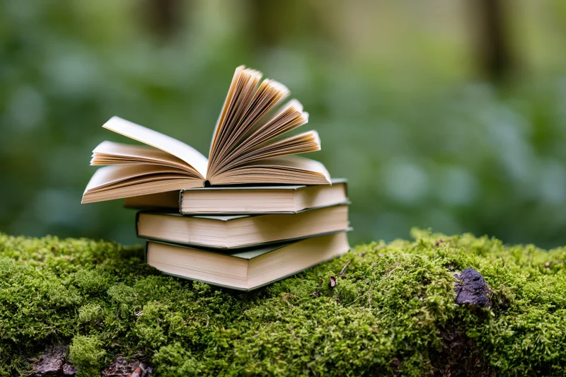 Stack of books with one open and fanned resting on mossy log in forest
