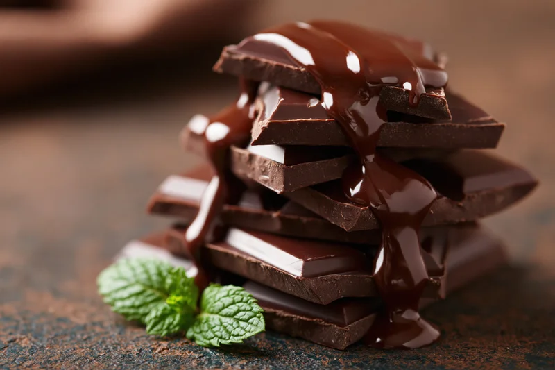 Broken dark chocolate pieces stacked high with chocolate sauce dripping over them, garnished with mint leaves on a textured surface