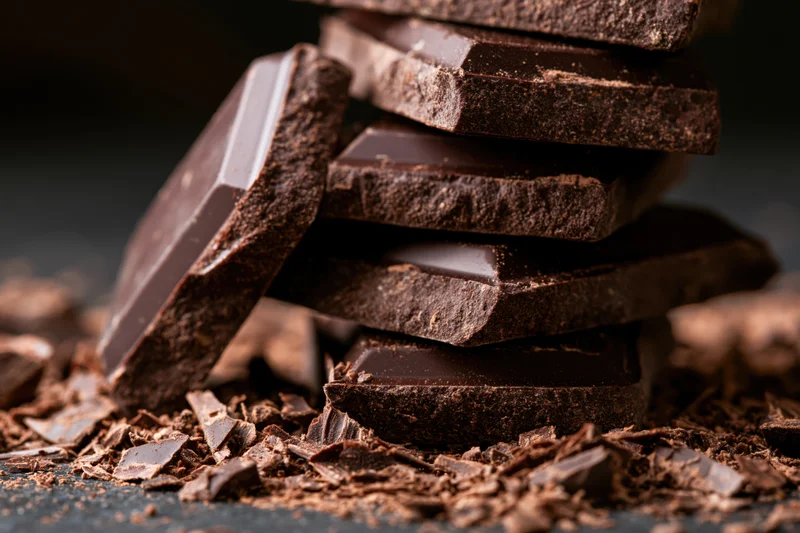 Broken dark chocolate slabs stacked on a dark surface with crumbs scattered around