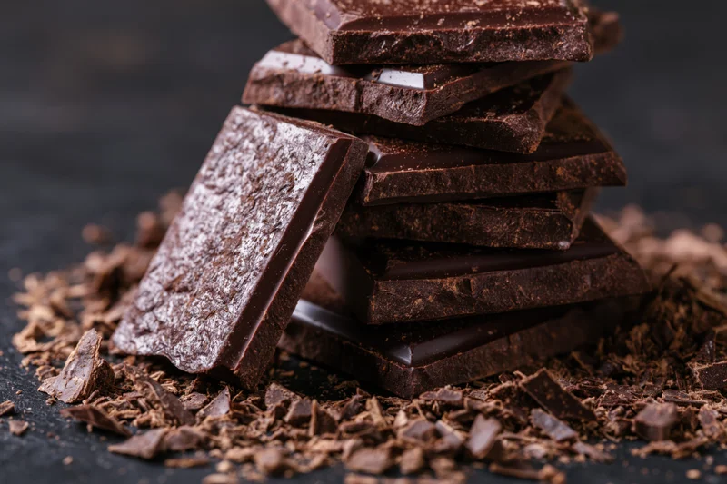 Broken dark chocolate slabs stacked on a dark surface surrounded by crumbs