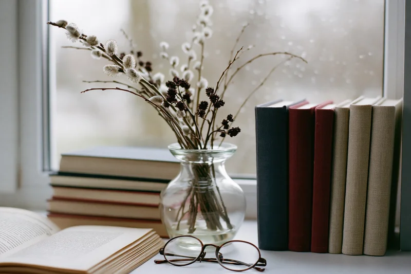 Stack of hardcover books beside a glass vase with pussywillow branches on a windowsill, with open book and reading glasses nearby