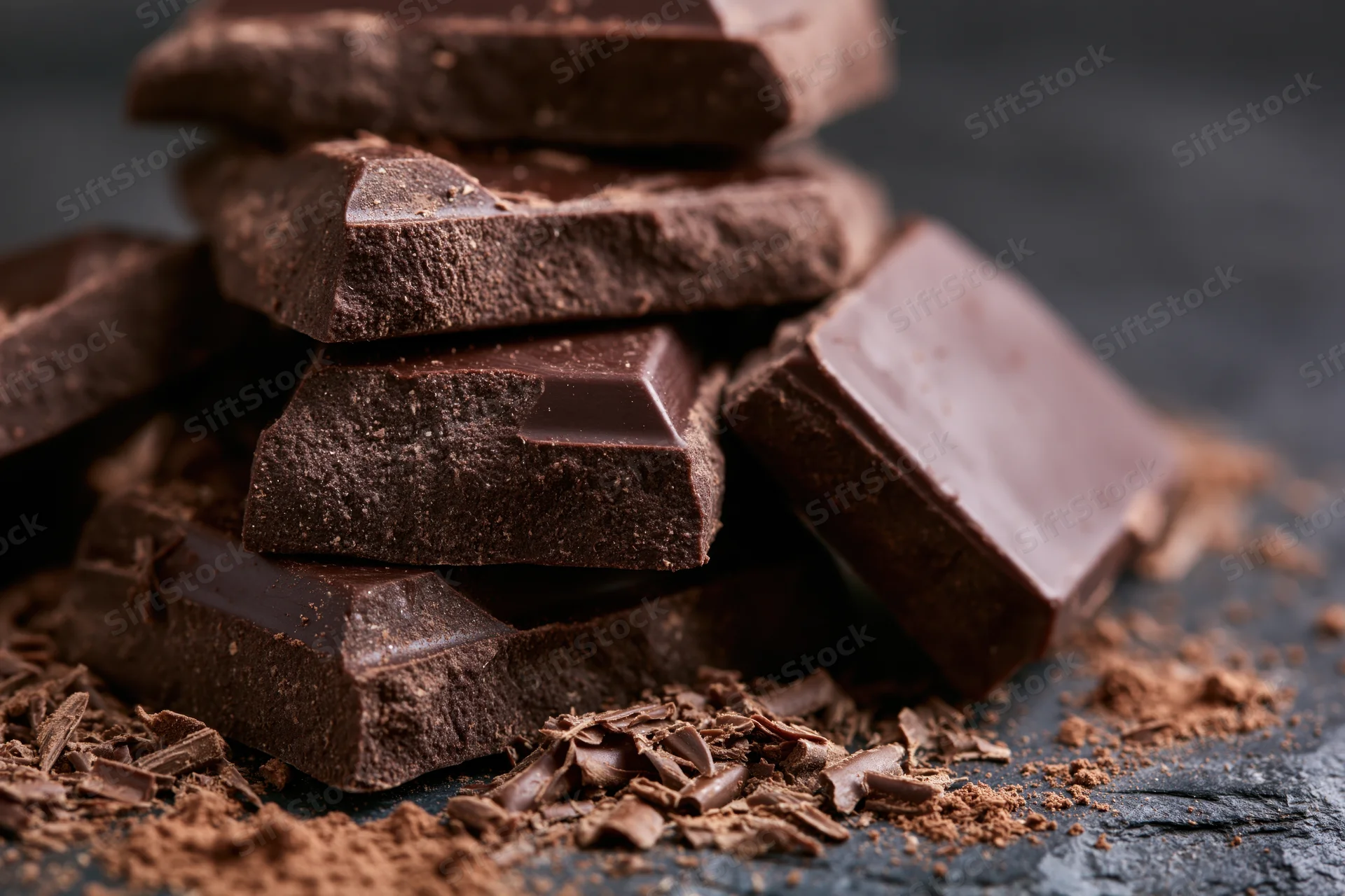 Broken dark chocolate chunks piled haphazardly on a dark slate surface with cocoa powder and crumbs scattered around