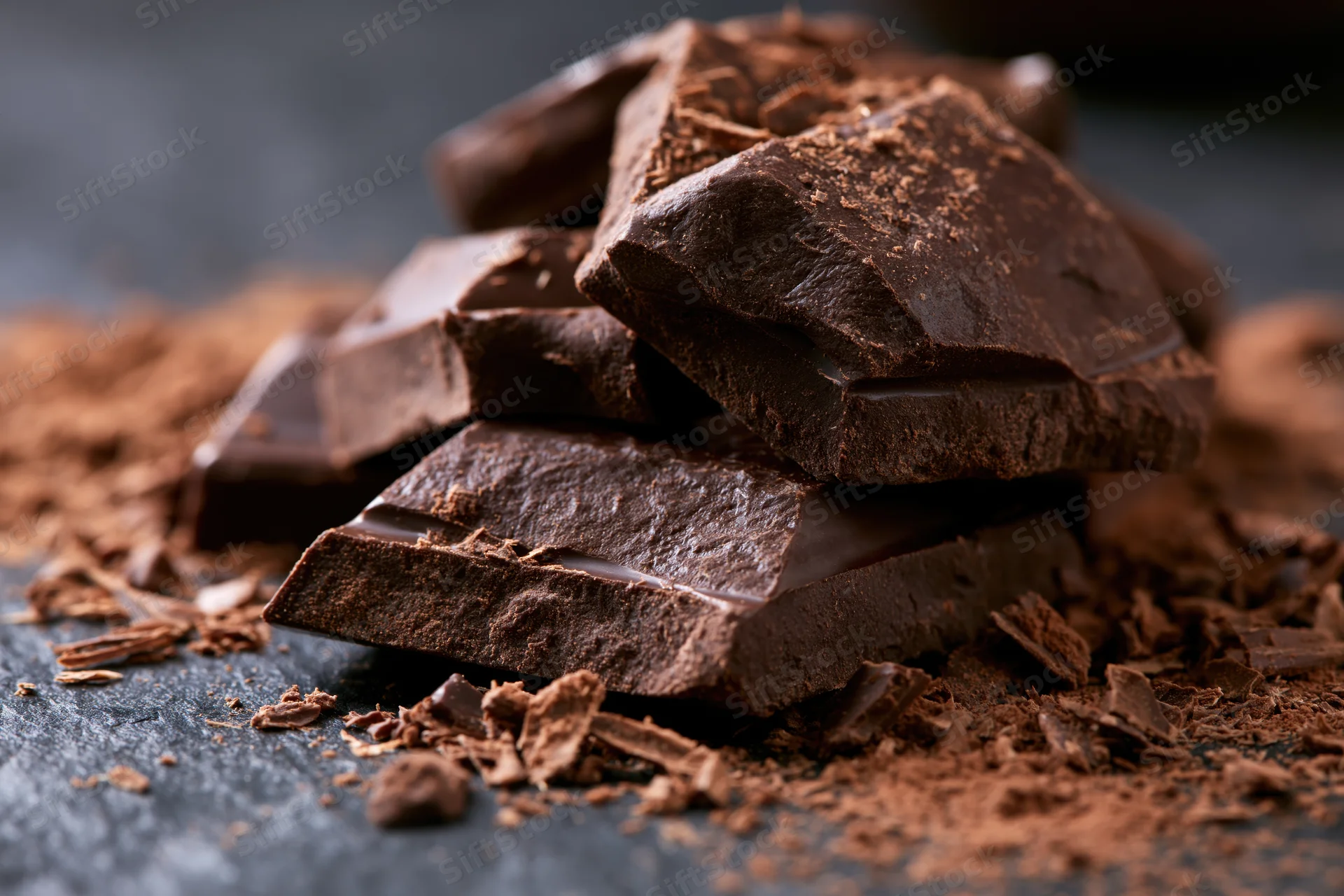 Broken dark chocolate chunks piled haphazardly on a dark surface with cocoa powder scattered around